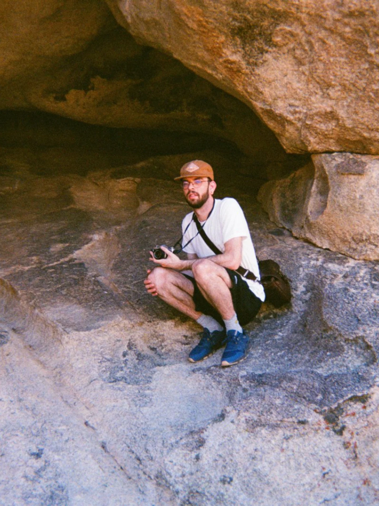 Ryan Cates in a cap and glasses, crouched with a camera in a sunlit rock alcove in Joshua Tree
